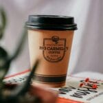 Kubek kawy z pokrywką na książce, w otoczeniu kwiatów i roślin