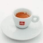 Kawa na czczo: czy picie espresso przed śniadaniem niszczy żołądek? Biała filiżanka espresso Illy ze spodkiem na jasnym tle